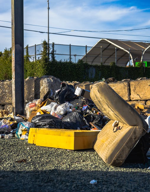 A large outdoor scene depicting a collection of overflowing rubbish bins and scattered waste materials on a paved urban sidewalk. The central focus is a grey mixed waste and paper bin with its lid ope