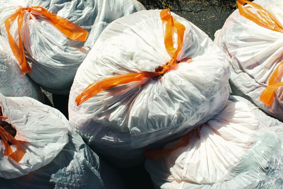 A group of white plastic rubbish sacks filled with waste materials, tightly sealed with orange plastic ties, are arranged outdoors on a paved surface. The sacks appear to contain various types of wast